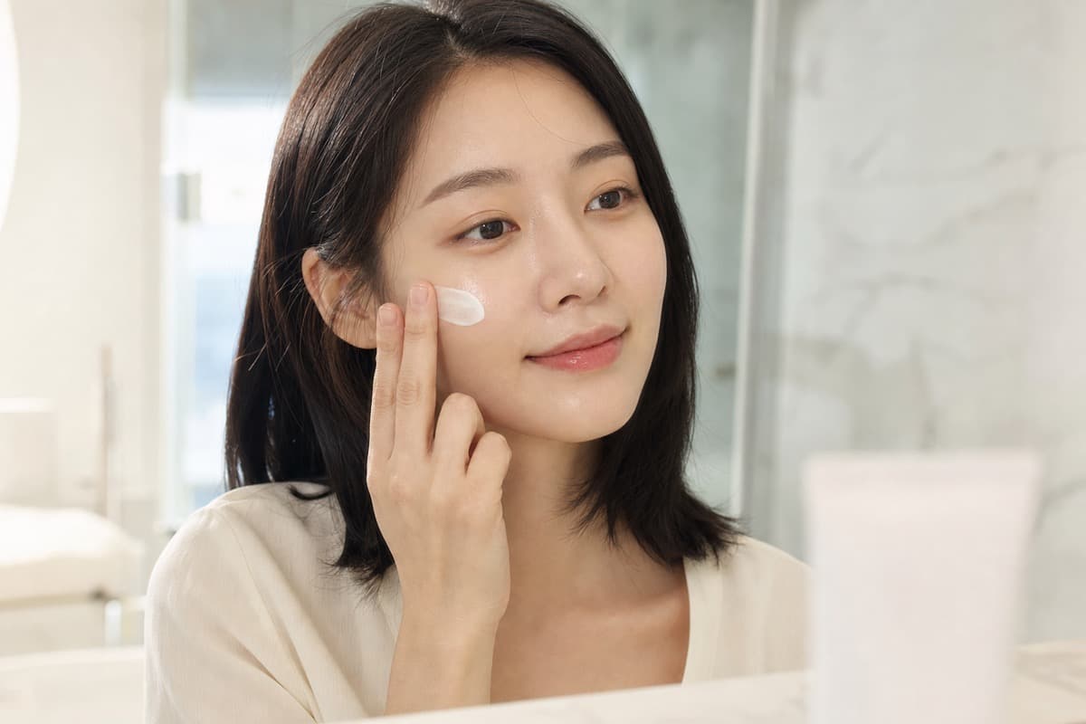Korean woman gently pressing a thin layer of unlabeled barrier cream onto her cheek in soft bathroom light