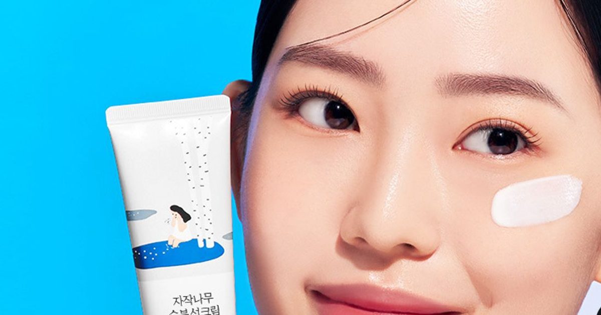 ROUND LAB Birch Juice Moisturizing Sunscreen white tube with a blue band held beside a model with a sunscreen swatch on her cheek